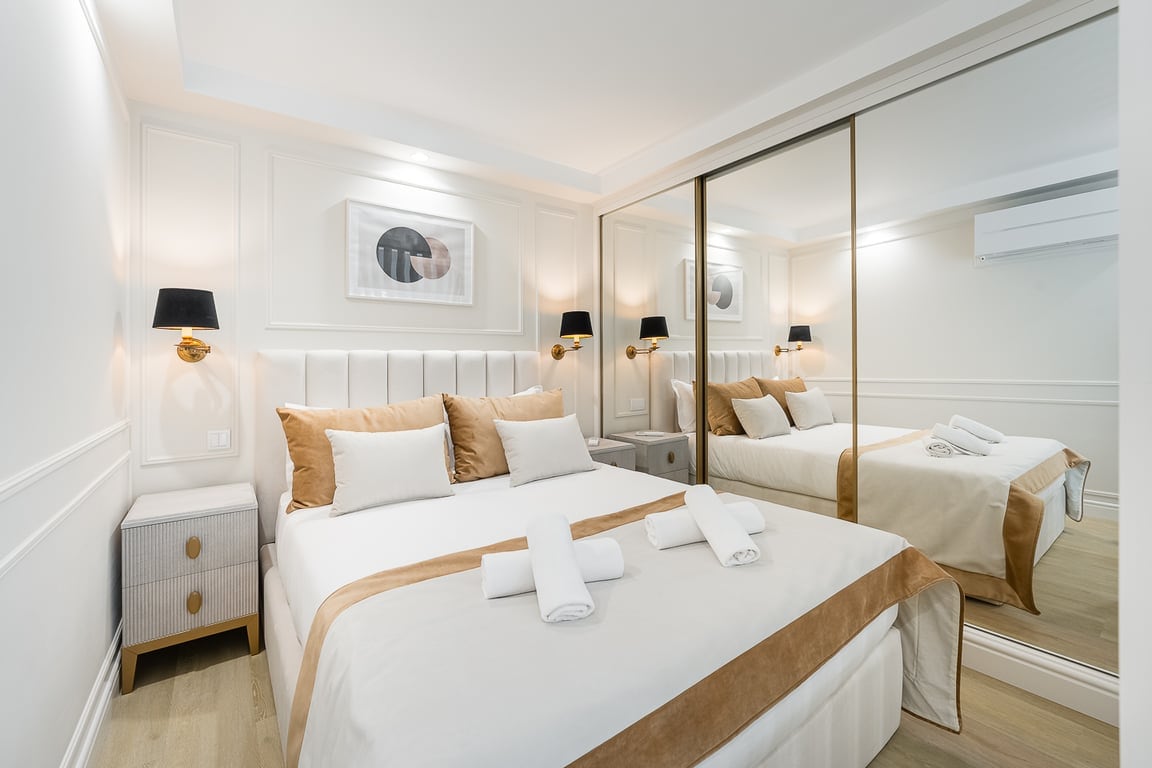A modern sleeping area features a spacious queen bed adorned with neutral bedding and decorative pillows. A mirrored wardrobe reflects the soft lighting. Stylish wall art and matching bedside lamps enhance the atmosphere, while a light wood floor adds warmth to the room.