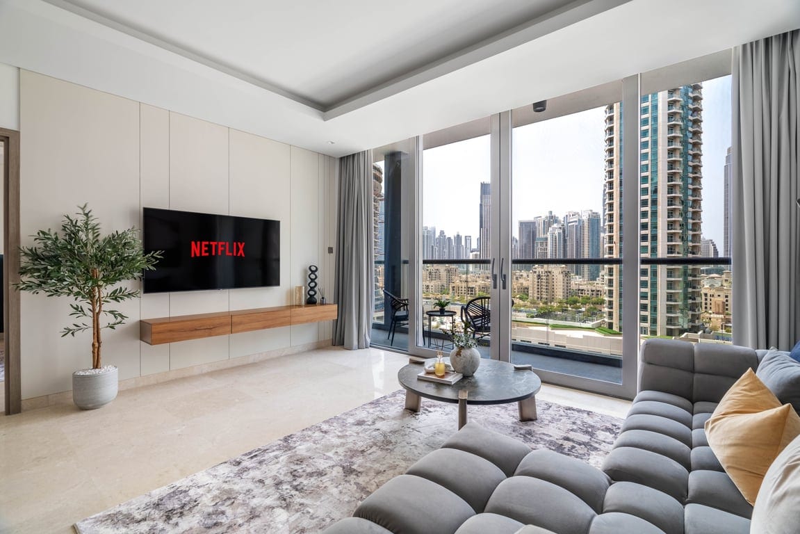 A bright living room welcomes guests with a large TV screen displaying Netflix. Floor-to-ceiling windows provide a view of the city skyline, while a comfortable sectional sofa is complemented by a small coffee table. A decorative plant adds a touch of greenery.