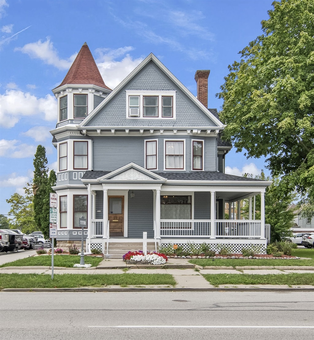 A historic three-story house with a pointed tower features a blue exterior with white trim and a welcoming porch. Lush greenery and flower beds enhance the front yard, while nearby streets reflect the vibrant atmosphere of downtown Springfield.