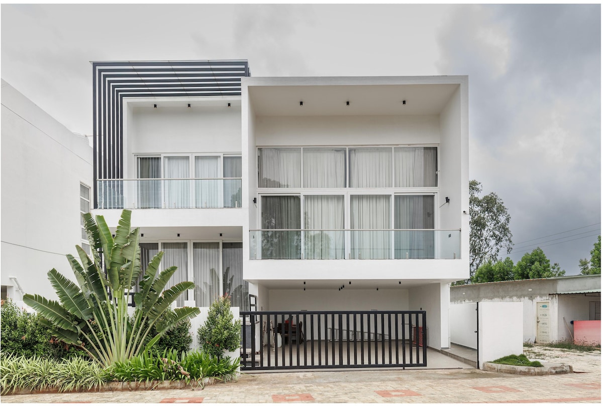 The villa's modern exterior features a sleek design with large glass windows and balconies. A spacious driveway is surrounded by landscaped greenery, including tall plants and manicured bushes, enhancing the inviting entrance. The structure is framed by a minimalist gate.