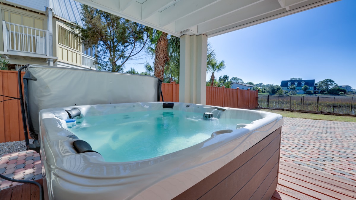 An outdoor hot tub is situated beneath an elevated patio, surrounded by a spacious deck. It features a clear, inviting water surface, with bubbles gently rising. Lush greenery and a clear sky are visible in the background, enhancing the serene outdoor setting.