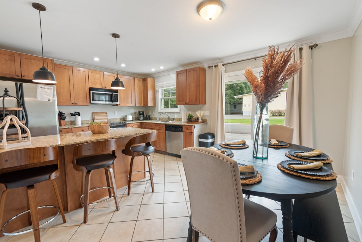 A modern kitchen is showcased with wooden cabinetry and stainless steel appliances. A spacious island counter with bar stools offers additional seating. A round black dining table is set for four, featuring woven placemats. Natural light floods the room through a window adorned with sheer curtains.