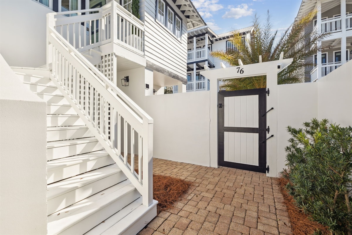 An inviting entrance is framed by a white stucco wall, with a wooden gate marked '76' visible in the center. A brick pathway leads to the gate, bordered by lush green plants and a palm tree, while a white staircase ascends on the left.