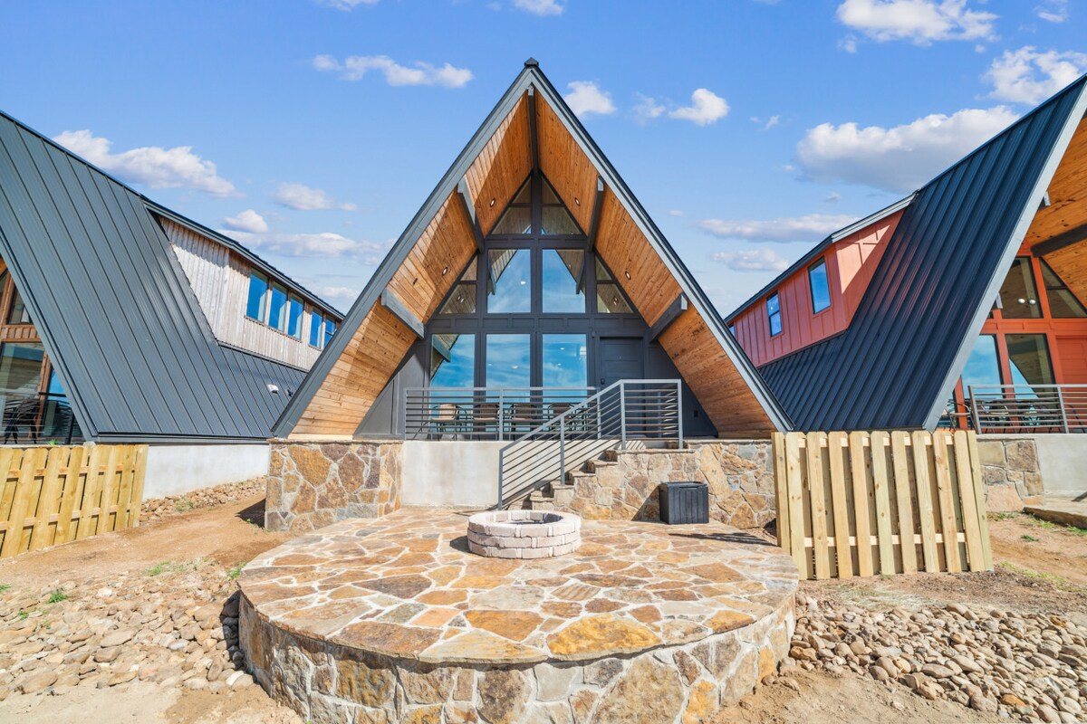 The A-frame cabin is framed by a rocky circular patio, featuring a fire pit at its center. Large windows in the front showcase the open interior, while the exterior design highlights a blend of wood and metal materials under a blue sky.