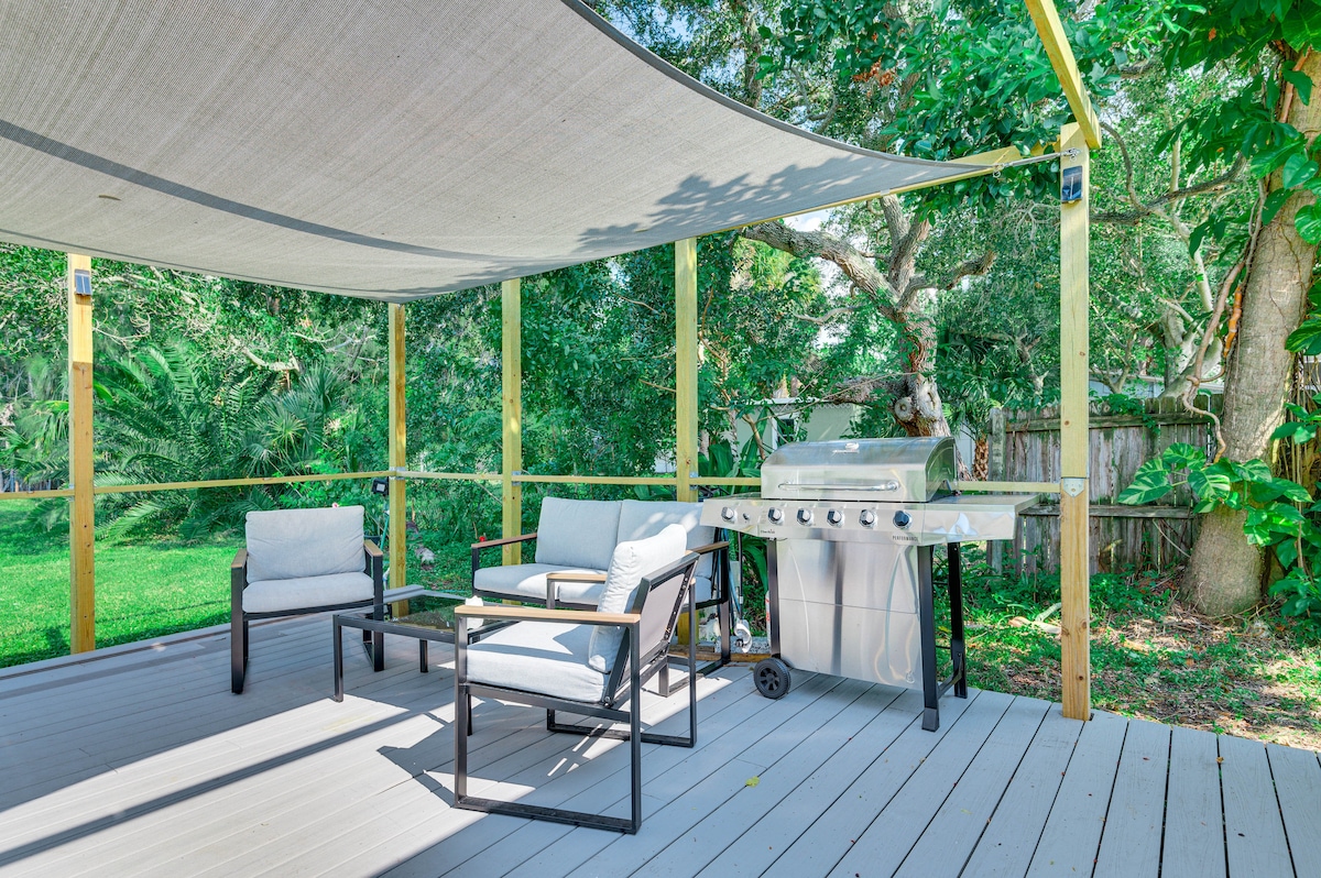 A furnished deck is shown with two lounge chairs and a propane grill. A sun shade sail provides protection from direct sunlight, while greenery surrounds the area, creating a private outdoor space for relaxation and barbecuing.