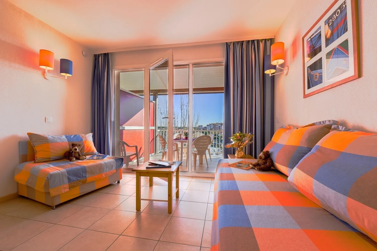 The living area features two colorful sofas with orange and blue patterns, complemented by warm lighting from wall-mounted lamps. Large glass doors open to the terrace, allowing natural light to fill the space. A wooden table sits in front, with decorative items placed on it.