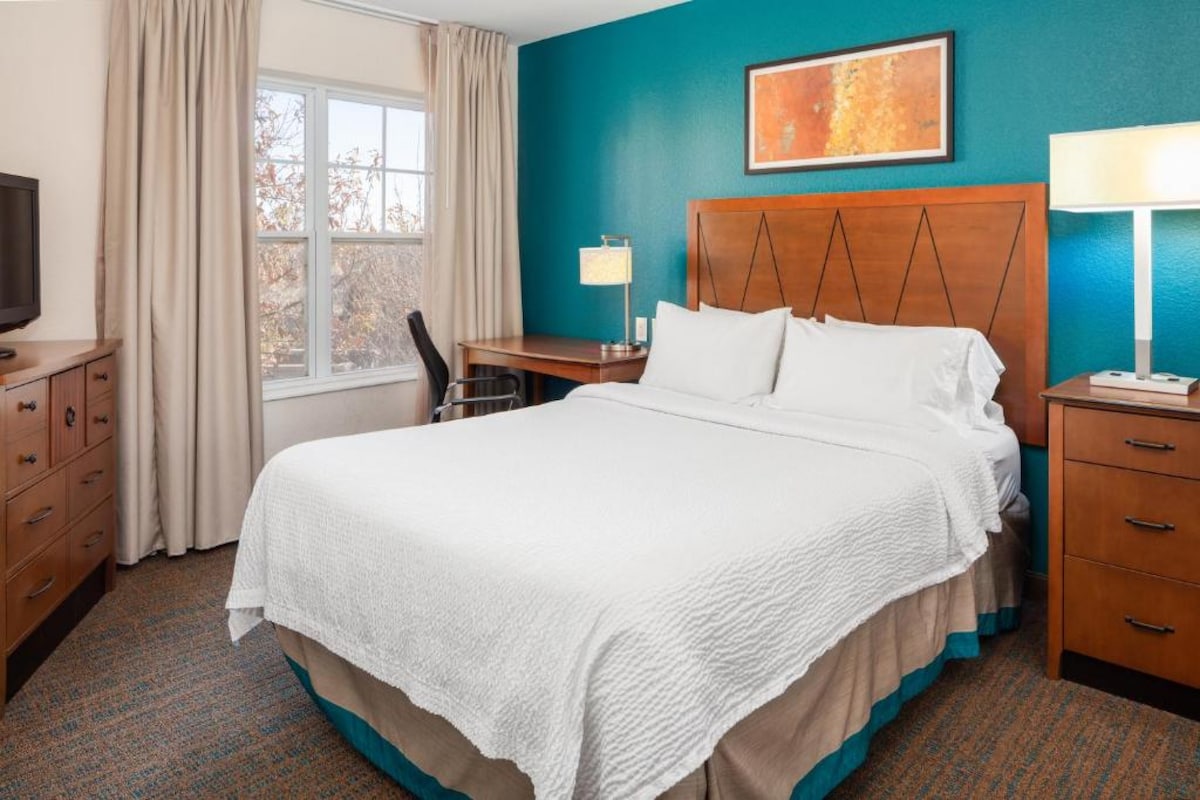 A well-appointed guest room features a spacious bed with crisp white linens against a backdrop of teal walls. A wooden desk and chair are positioned near the window, which allows natural light to brighten the room. A flat-screen TV is mounted on the adjacent dresser.