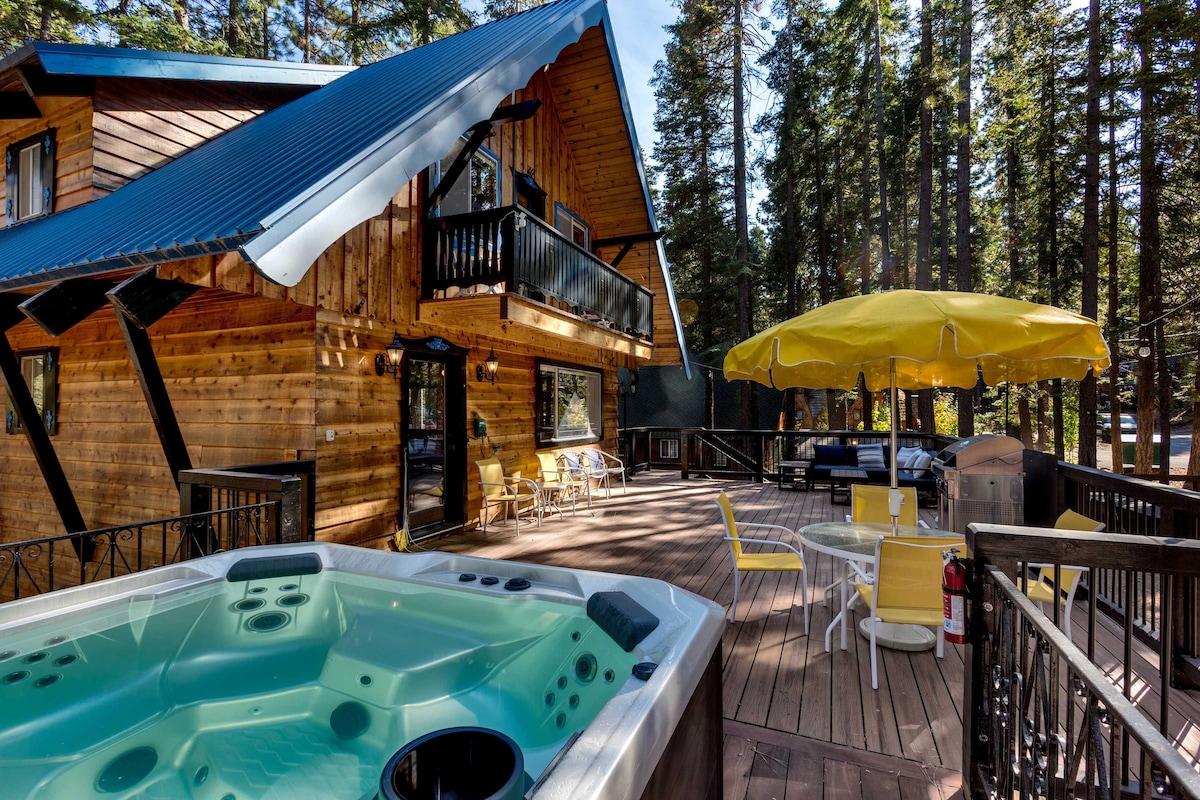 An expansive wooden deck surrounds a modern hot tub, providing a relaxing outdoor space. Yellow patio furniture is arranged under a bright umbrella, complemented by additional seating options. Towering trees create a natural backdrop, enhancing the serene ambiance of this getaway.