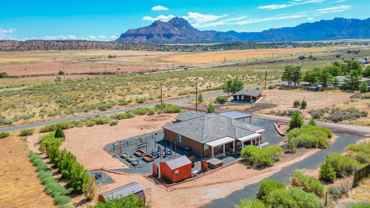 The exterior view showcases a spacious retreat surrounded by open fields and distant mountains. A paved driveway leads to the home, which is situated within a well-maintained outdoor area featuring a variety of plants, outdoor seating, and accessed by wide dirt paths.