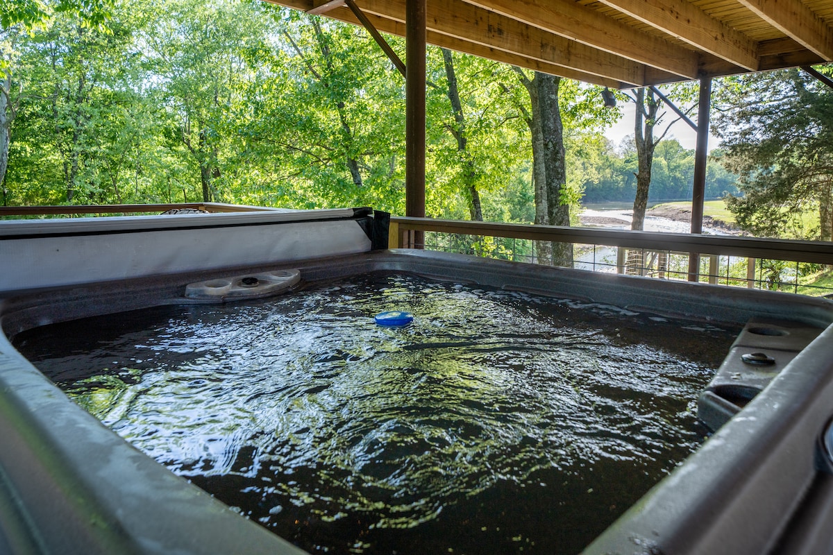 A spacious hot tub is situated under a wooden deck, offering views of lush trees and the nearby river. The water surface gently ripples, reflecting the natural light filtering through the surrounding foliage.