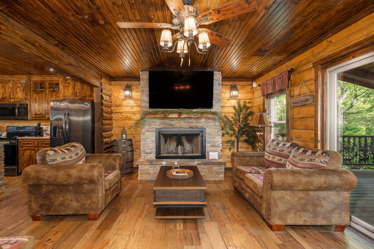 A warm living area features a stone fireplace and a large flat-screen TV mounted above it. Two comfortable sofas with decorative cushions and a wooden coffee table create an inviting space. Natural wood accents and lighting enhance the rustic charm.
