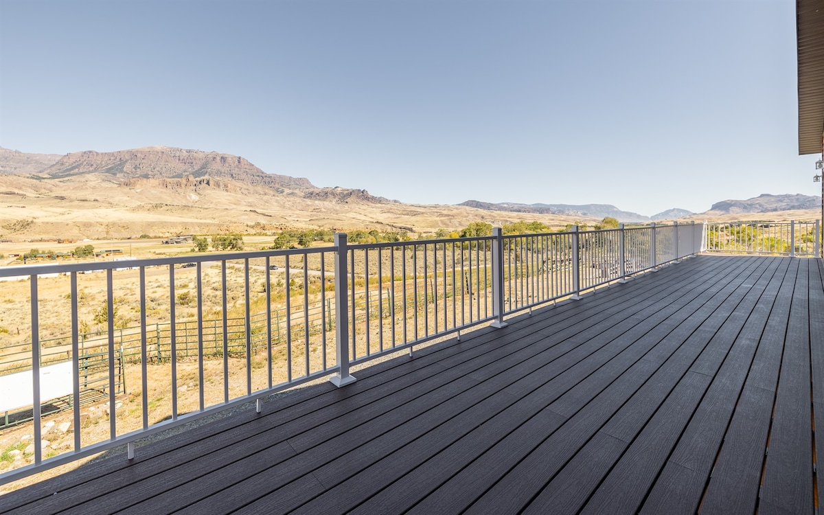 A spacious deck extends outward, surrounded by a sleek metal railing. The open space offers unobstructed views of the vast, dry landscape and distant mountains, showcasing a serene natural environment under a clear blue sky.