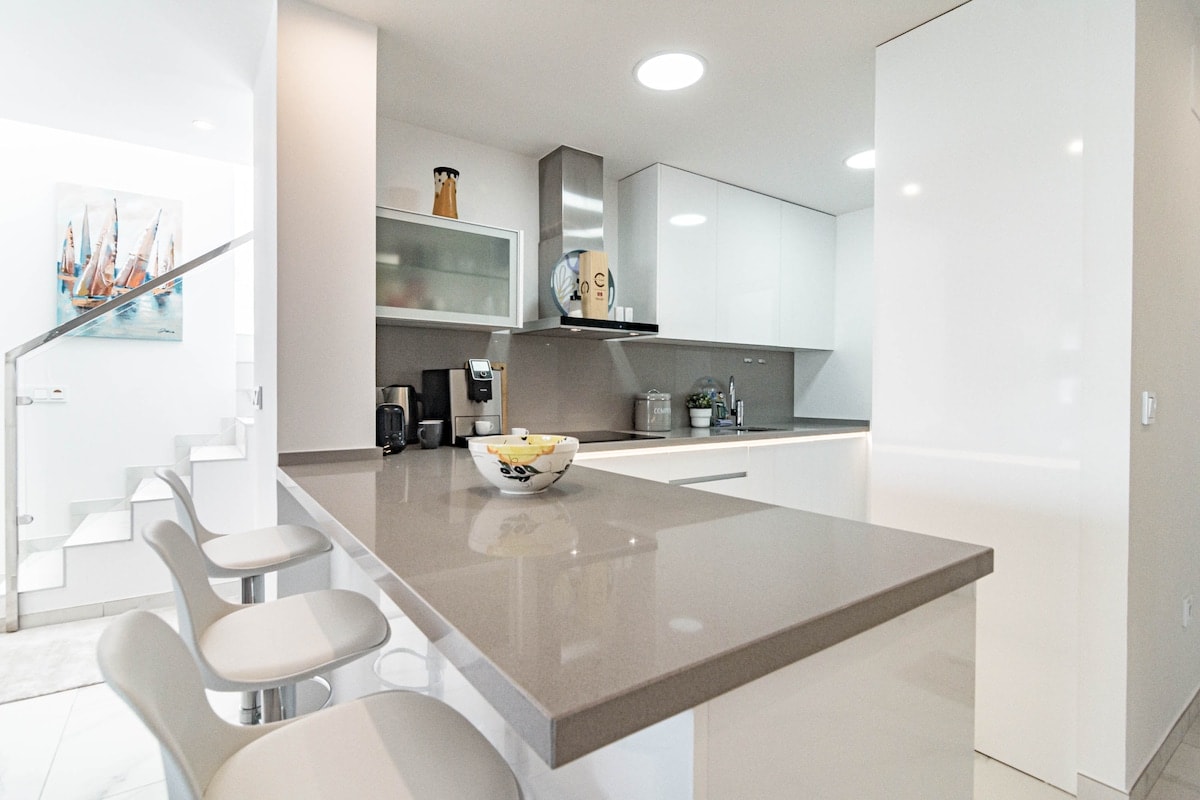 The modern kitchen features sleek white cabinetry and a spacious grey countertop. Barstools are positioned at the counter, providing seating for three. A painting of sailing boats adds a touch of color to the room, while natural light illuminates the area.