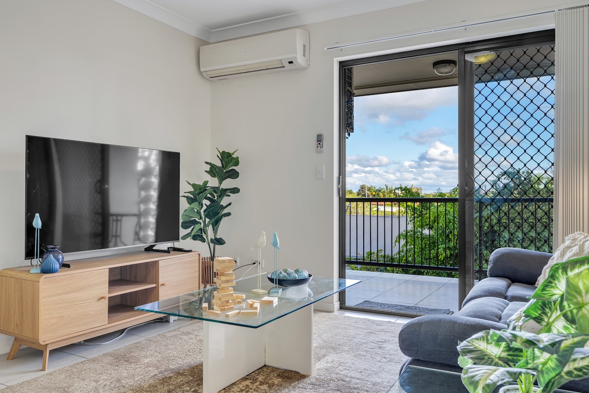A bright living area is presented, featuring a comfortable couch and a sleek glass coffee table. A television is mounted on a light wooden TV unit. Natural light enters through large sliding doors that lead to a private balcony, with greenery visible beyond.