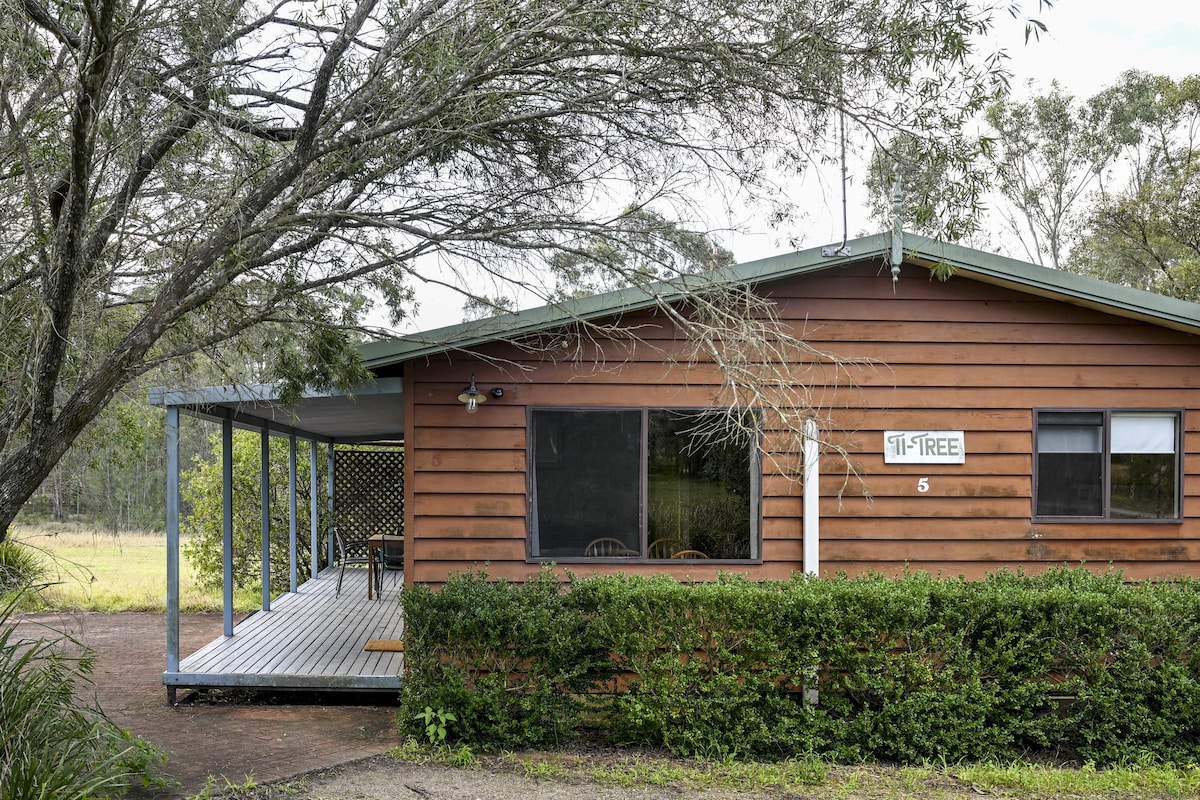 A rustic cabin is set against a natural backdrop, featuring earthy wooden siding and a covered patio area. Seating is visible on the deck, with a large window offering views of the surrounding greenery. Lush hedges frame the entrance to create a sense of privacy.