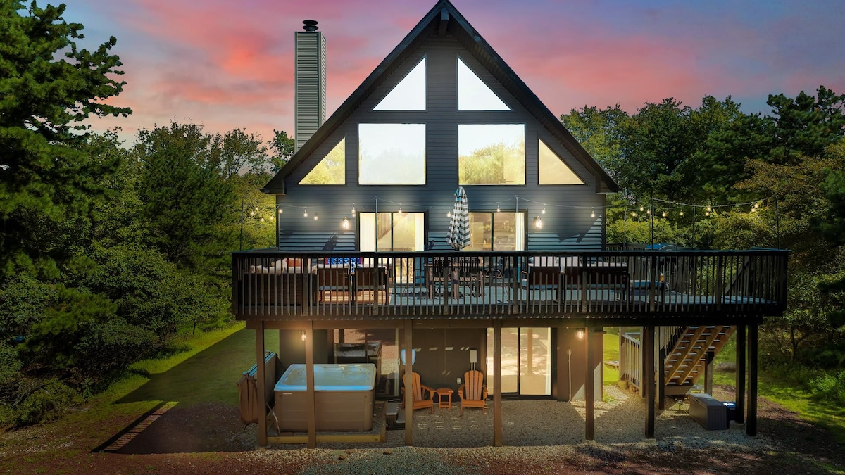 The cabin exterior showcases a modern design with a striking A-frame structure. Large glass windows invite natural light, while a spacious deck features seating and a hot tub, surrounded by trees. Soft evening light casts a warm glow over the peaceful setting.