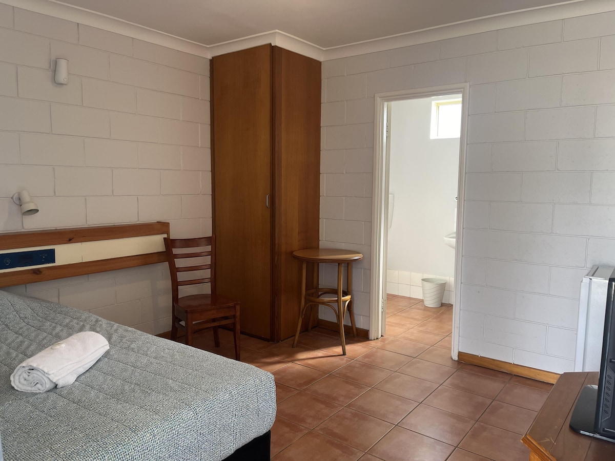 A studio room is depicted, featuring a king single bed with a patterned cover. A wooden chair and small table occupy the space, complemented by a wardrobe. Natural light enters through a window, illuminating the tile floor and providing a view of the ensuite bathroom.