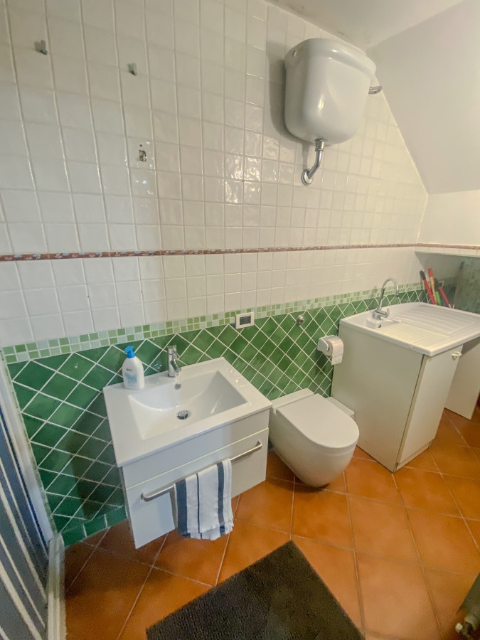 A functional bathroom is presented, featuring a white toilet and a compact sink with a mirror above. Green and white tiles create a patterned backsplash, while a washing machine is positioned beside the sink. A bath mat is laid on the tiled floor.