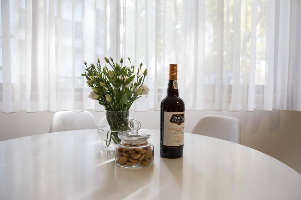 A round table is set elegantly with a transparent glass vase filled with white flowers and a bottle of port wine. A glass jar containing nuts rests on the table, bathed in natural light that filters through sheer white curtains.