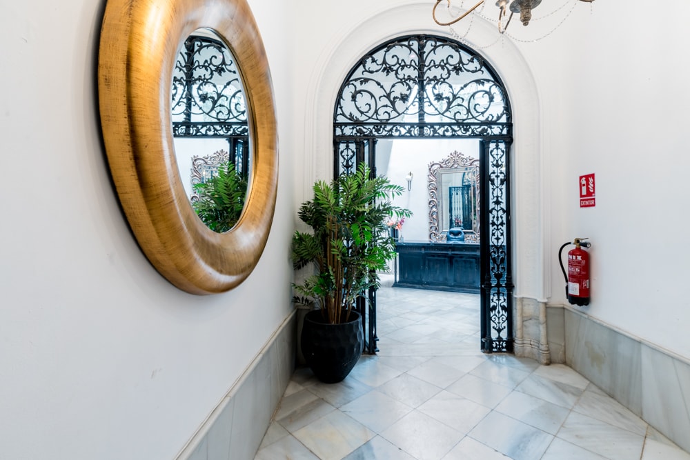 An inviting corridor features a large, round golden mirror reflecting the ornate entrance area. An elegant black metal gate leads to a landscaped courtyard, while a potted plant adds a touch of greenery beside the marble flooring.