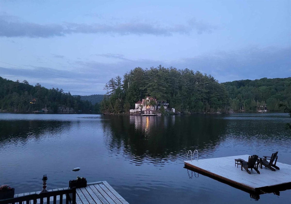 A serene view of Lac St. Pierre is captured at dusk, featuring calm waters reflecting the surrounding greenery. In the distance, an isolated island is visible, surrounded by tall trees. A wooden dock extends into the lake, leading to a swimming ladder.