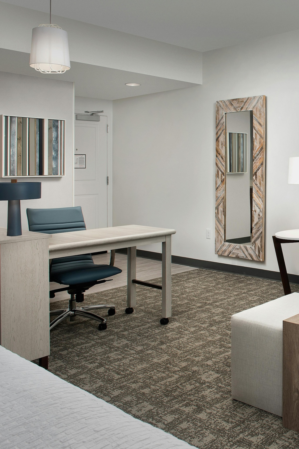 A well-organized room features a workspace with a desk and rolling chair, complemented by a modern lamp. A full-length mirror reflects the light from the nearby window. Textured carpet covers the floor, while light-colored furnishings provide a bright and airy feel.