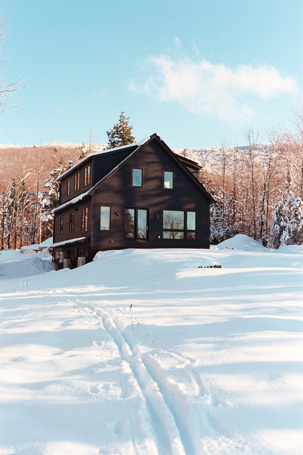 The modern black house is set against a backdrop of a winter landscape, with snow-covered ground reflecting the sunlight. Large windows are visible, allowing natural light to fill the interior. Snow-draped trees and rolling hills create a serene winter atmosphere.