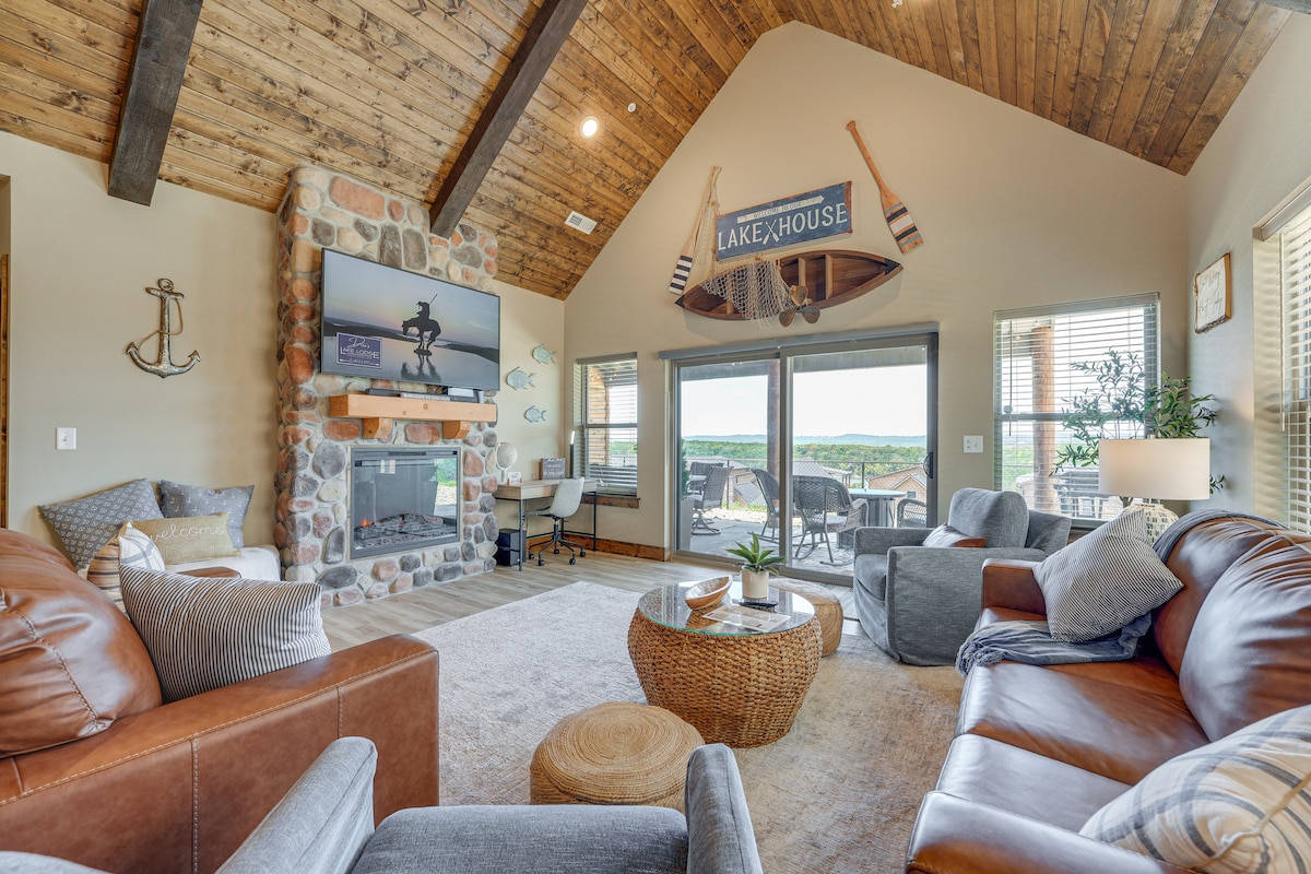 The living room features a high, vaulted ceiling with exposed wooden beams, creating an open atmosphere. A stone fireplace is central, flanked by a large Smart TV. Comfortable seating includes a leather sofa and armchairs, with large windows providing views of the surrounding landscape.