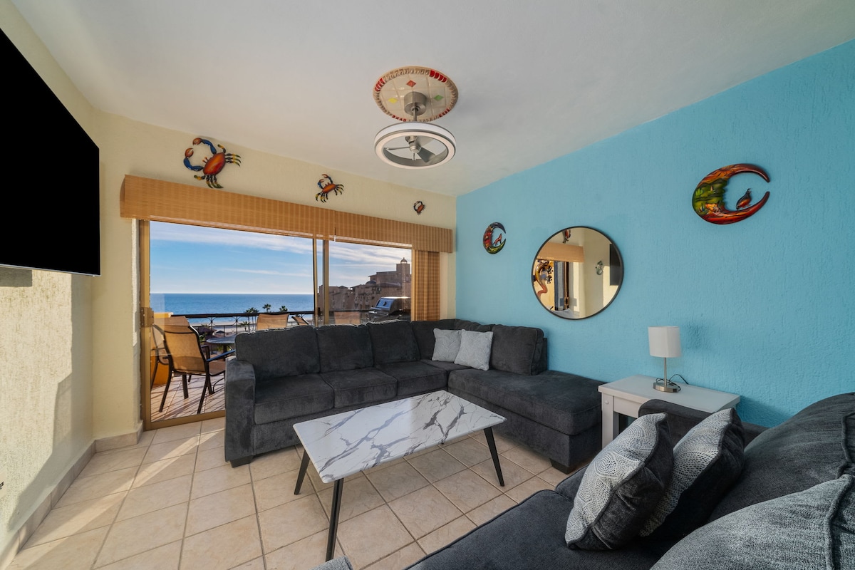 A spacious living area features a large grey sectional sofa positioned around a coffee table with a marble finish. Two circular mirrors reflect natural light, while artwork depicting crabs adorns the walls. The sliding doors open to a view of the sparkling sea and outdoor space.