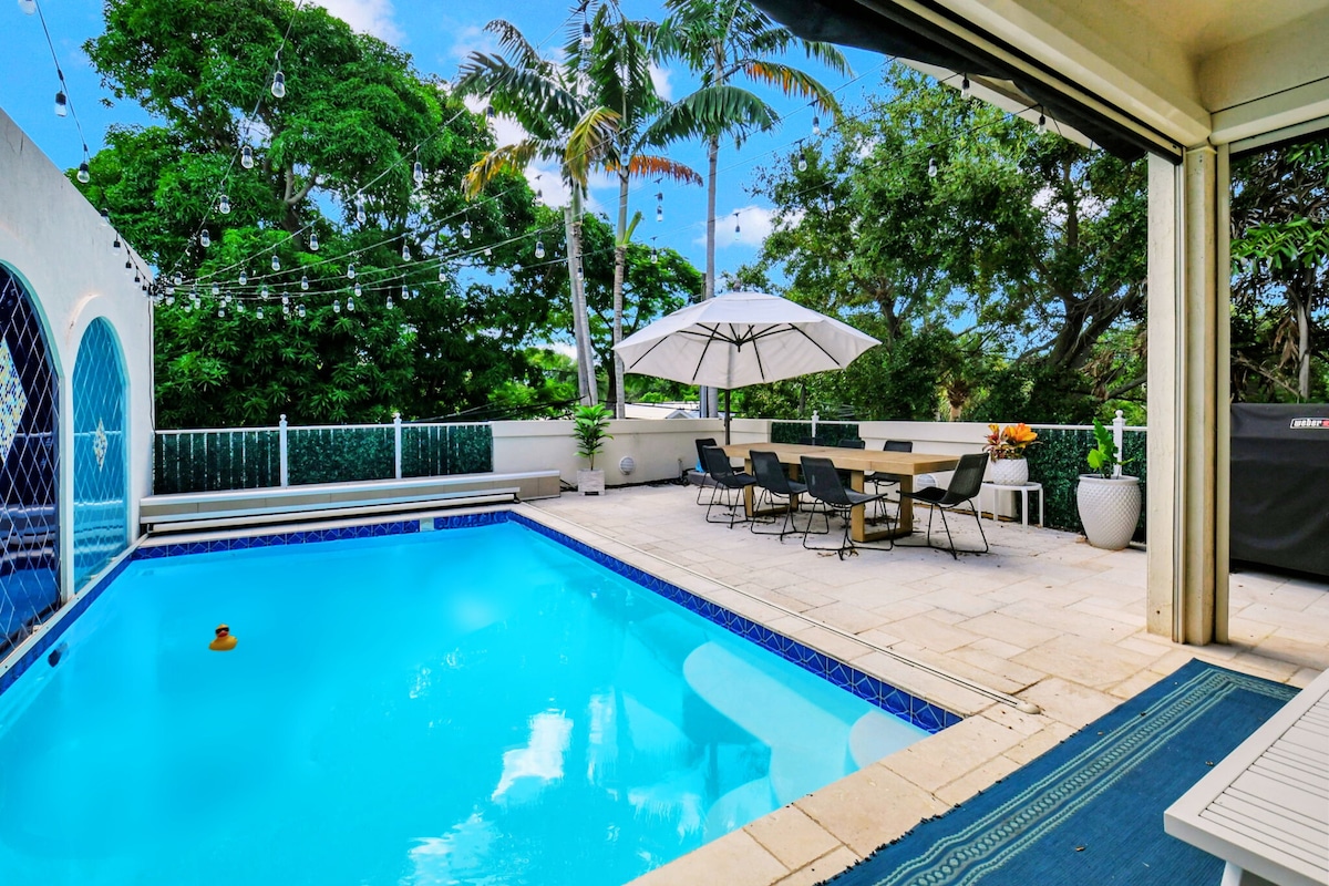 A private saltwater pool is surrounded by a spacious patio area with multiple seating options. An umbrella provides shade over a large dining table, while string lights add a subtle ambiance. Lush greenery frames the backyard, enhancing the serene outdoor setting.