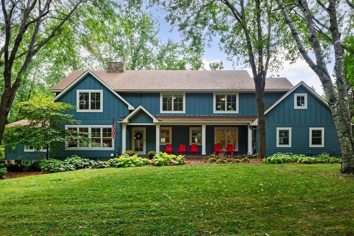 The exterior of a two-story blue house is surrounded by lush greenery and mature trees. A well-maintained front yard features a grassy area and a pathway leading to the entrance. Red chairs are positioned on the porch, inviting a relaxed atmosphere.