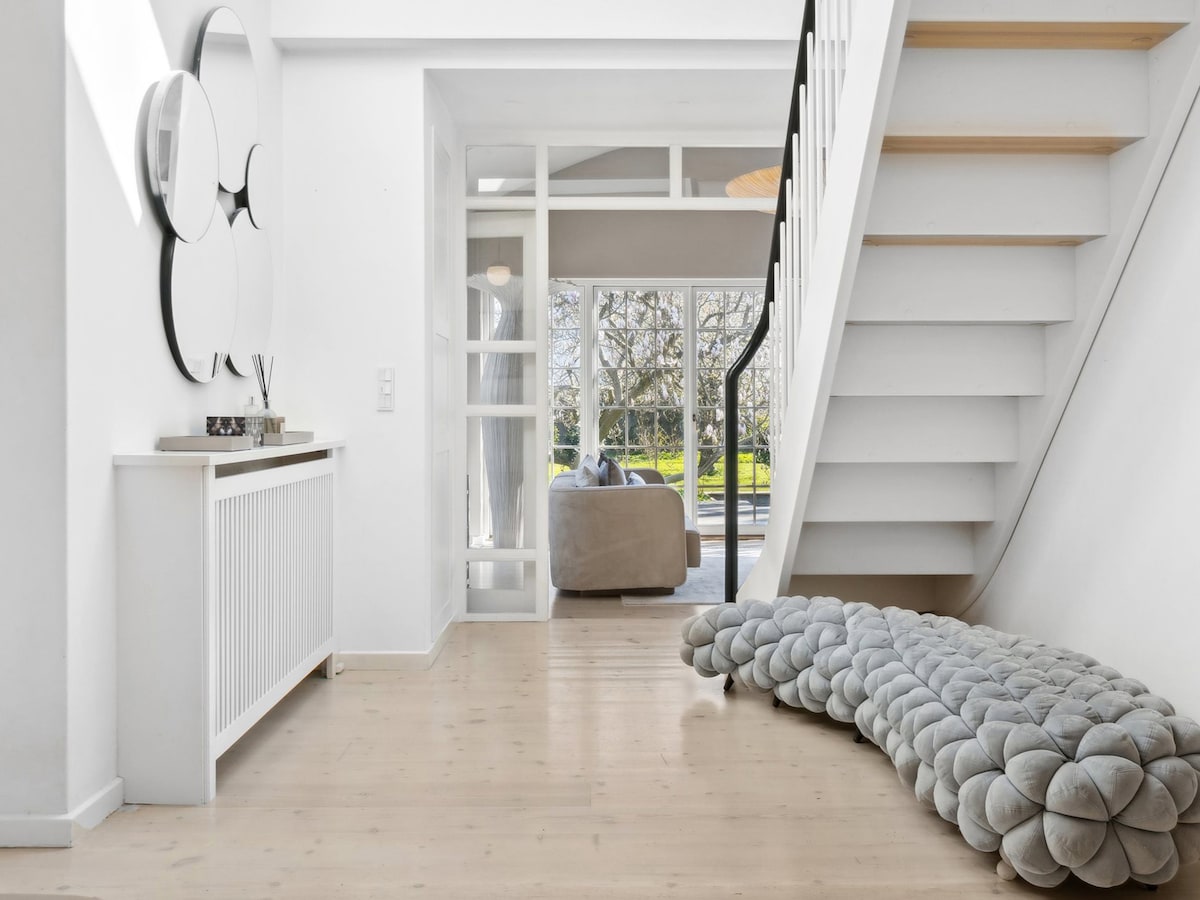 A bright entrance area is characterized by light-colored wooden flooring. A sculptural seating piece in a soft, curved design adds a contemporary touch. A staircase leads to the upper levels, with glass panels framing the view of the garden visible through large windows.