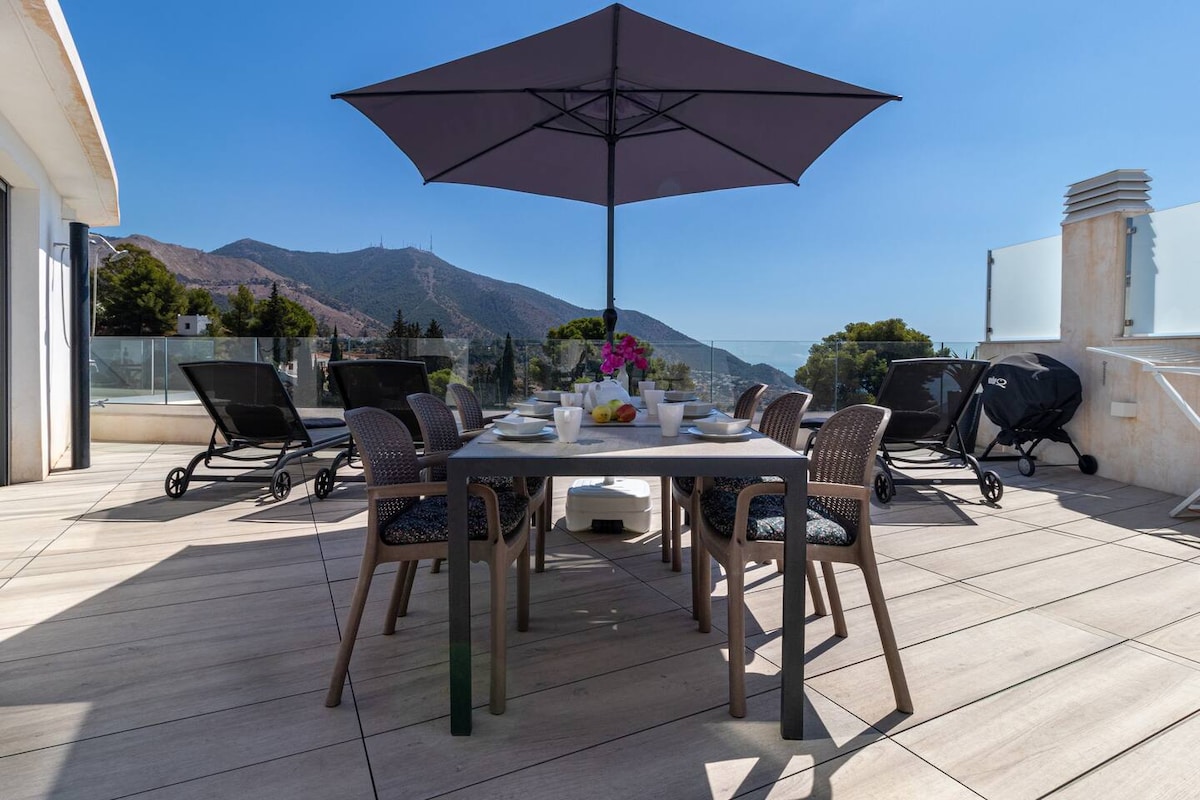 A spacious terrace is presented with a dining table set for six under a large umbrella. Sun loungers are arranged around the area. Lush greenery and gently sloping mountains are visible in the background, enhancing the outdoor experience.