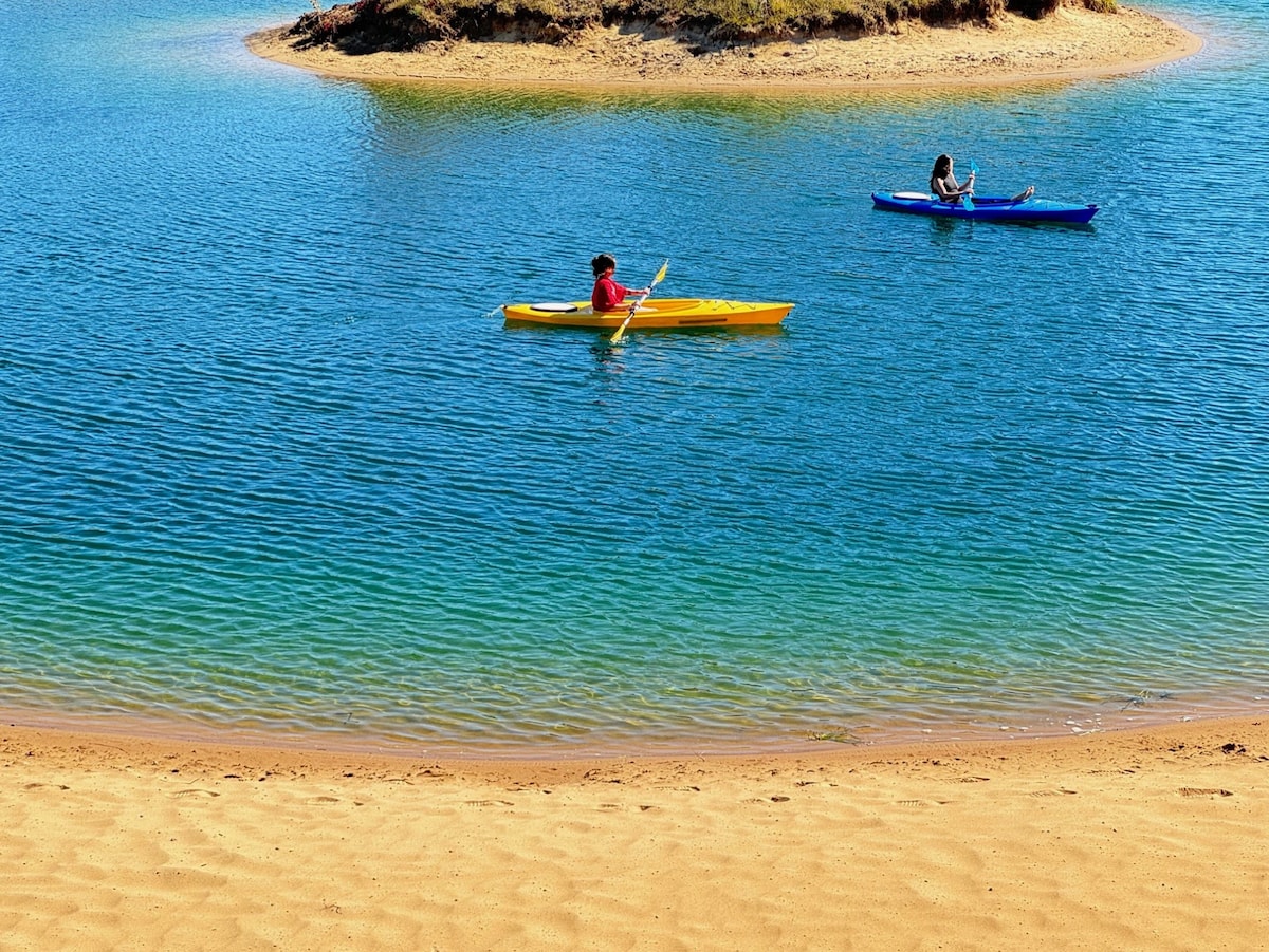 Two kayakers paddle across clear turquoise waters near a sandy beach. A small, grassy island is visible in the background, enhancing the natural setting. The area offers a serene atmosphere, inviting guests to engage in water activities at the lakefront.