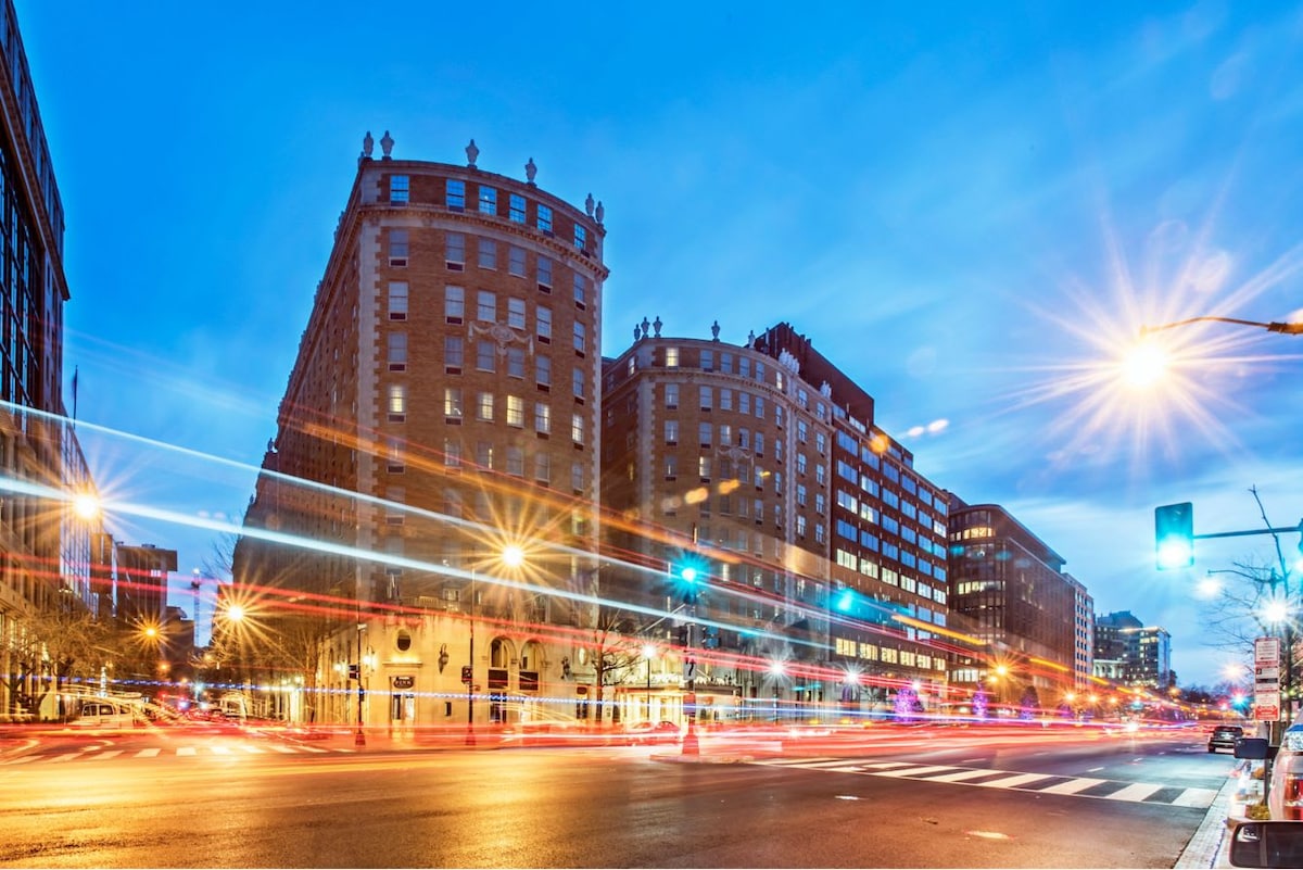 The exterior of a historic building is captured at dusk, illuminated by streetlights and vehicle traffic. Architectural details are visible on the curved façade, while nearby roads and bustling city life create a vibrant atmosphere in downtown Washington, D.C.