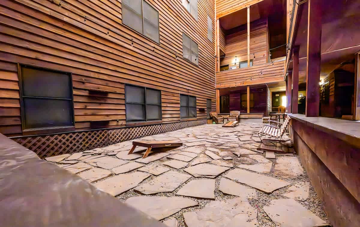 A communal outdoor space is surrounded by wooden structures, featuring a stone-paved courtyard. Several lounge chairs are positioned on the stone surface, providing areas for relaxation. Soft lighting is evident in the background, creating a gentle ambiance for evening gatherings.