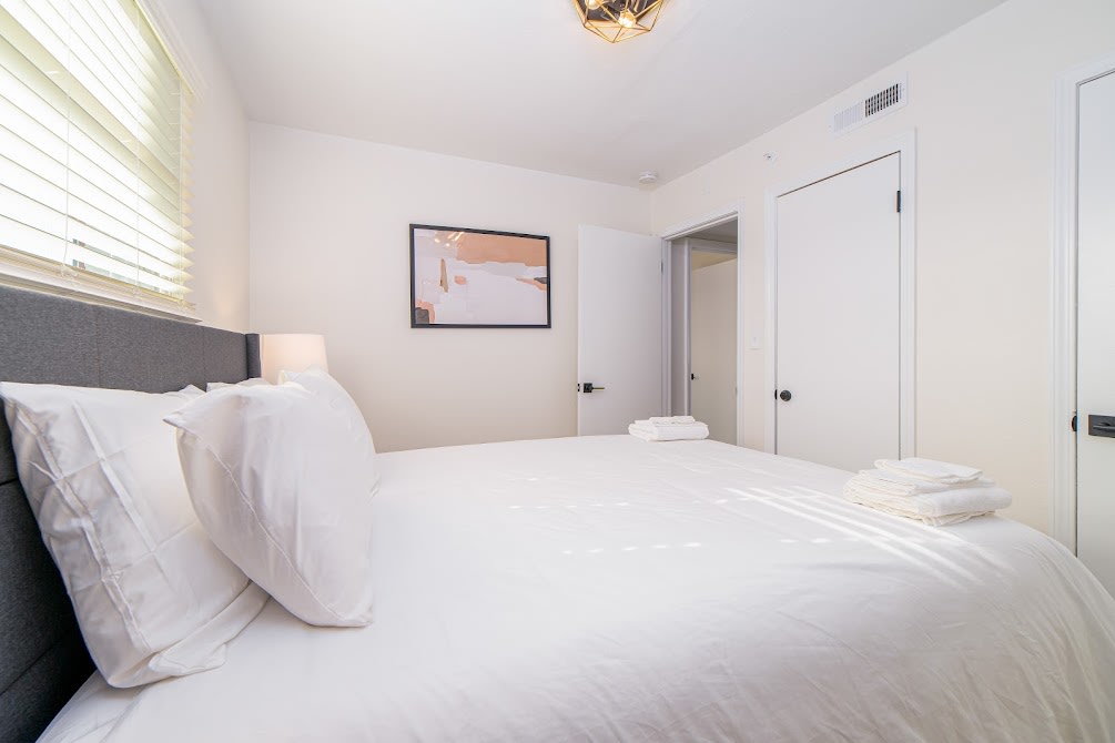 A bright bedroom features a spacious bed with crisp white linens and neatly arranged towels. Soft natural light filters through a window with blinds. A piece of artwork adorns the wall, with a closet and a bathroom door visible in the background.