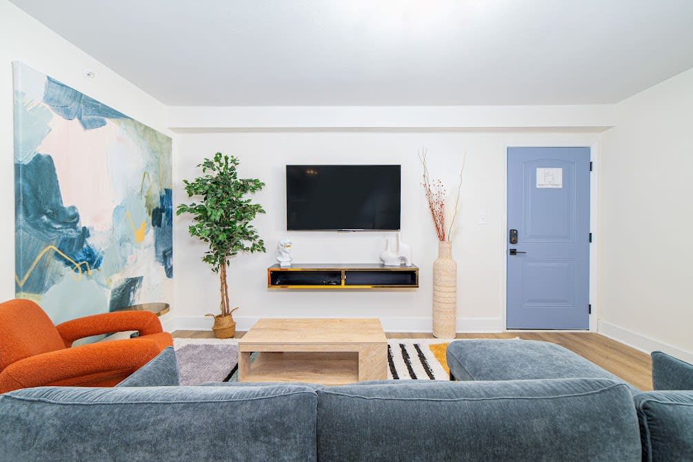 The living room features a modern design with a large gray couch and an orange armchair. A wooden coffee table sits centrally, with a vibrant artwork displayed on the wall. A flat-screen TV is mounted above an entertainment unit, and a potted plant adds greenery.