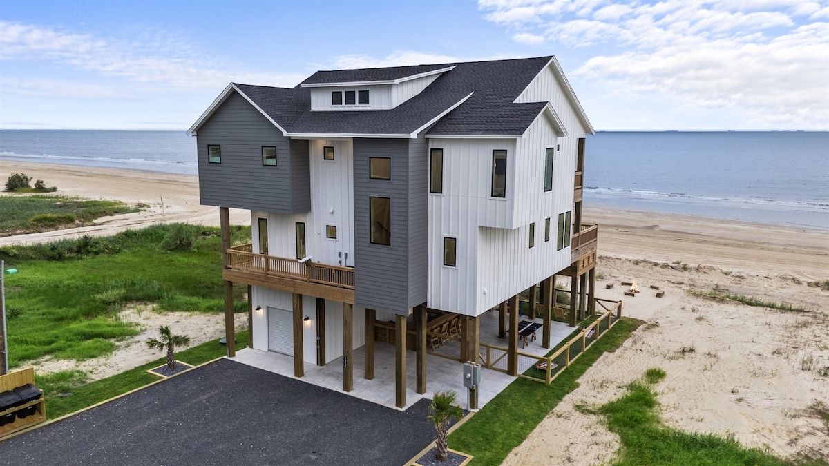 A two-story beachfront retreat is presented, showcasing a contemporary architectural design with a mix of white and gray siding. Elevated above ground, spacious decks are visible, offering direct views of the sandy beach and ocean. The surrounding landscape features greenery and a parking area.