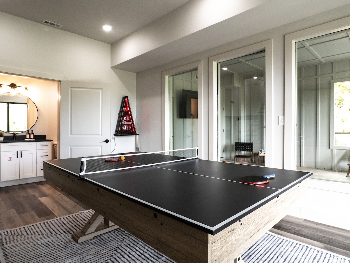 A game room features a sleek ping pong table with paddles and balls ready for play. Large glass doors provide natural light and access to an outdoor area. The room is completed with modern decor and a striped area rug, enhancing the inviting atmosphere.