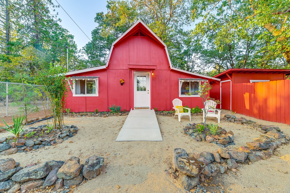 The exterior of the red cottage features a well-maintained walkway leading to the entrance. Two white chairs are positioned on the porch, surrounded by a landscaped area with rocks and low greenery, creating a welcoming entryway.