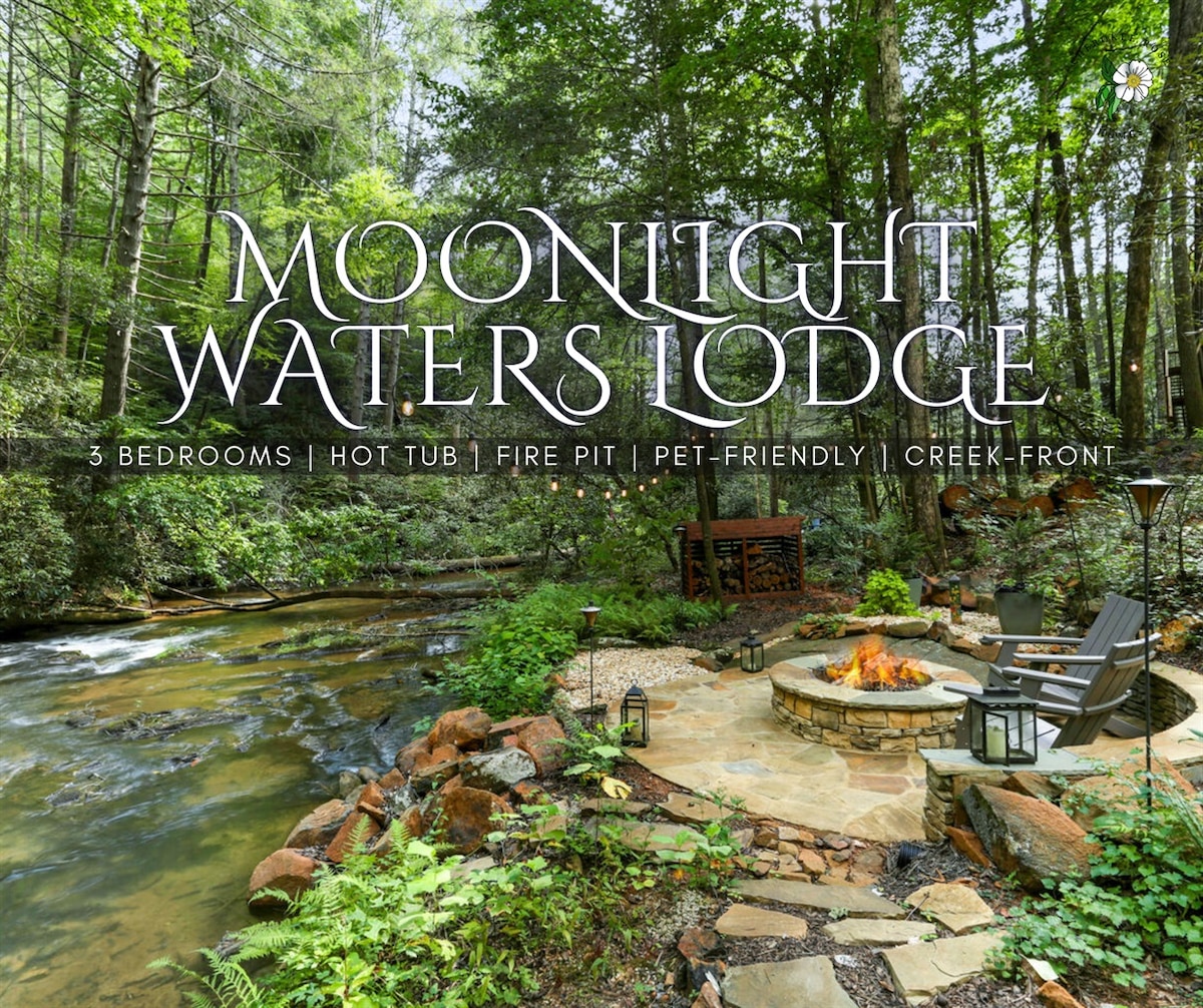 An outdoor seating area features a stone patio overlooking a serene creek, surrounded by lush trees. The fire pit is arranged with seating around it, creating a cozy atmosphere. A hot tub is visible nearby, inviting relaxation in nature's embrace.