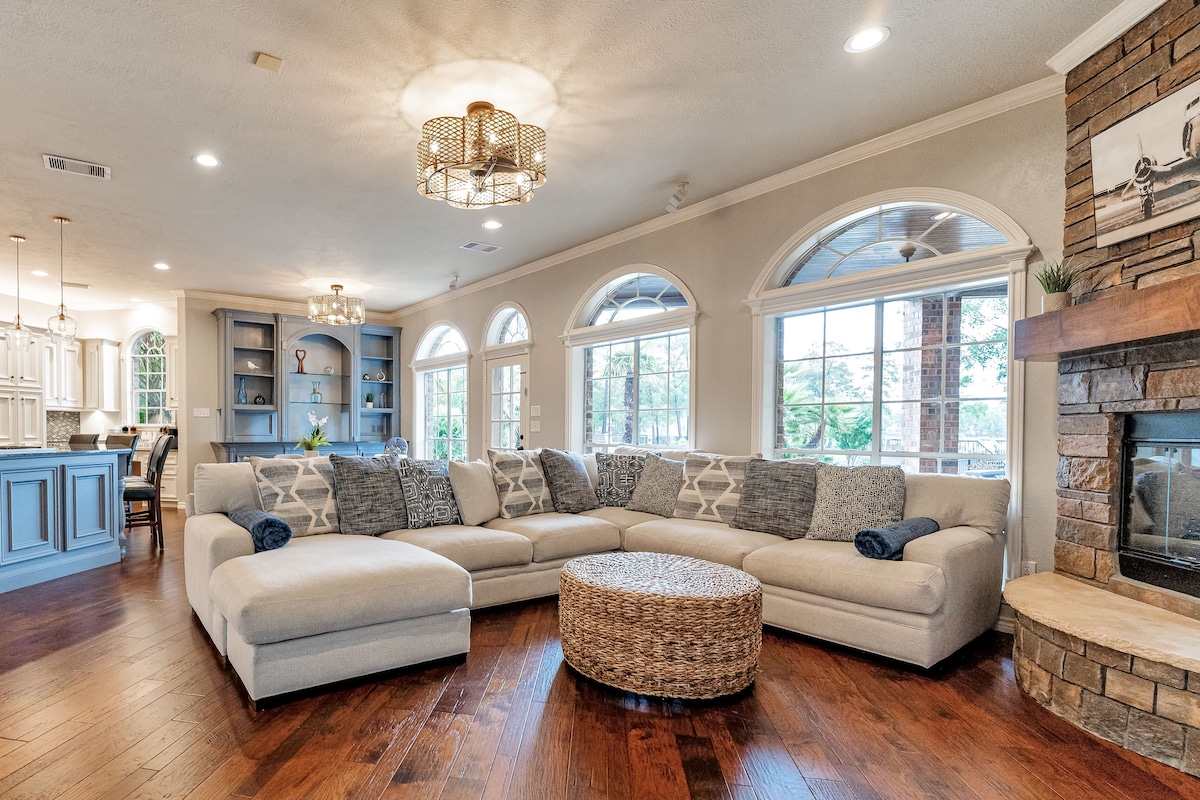 A spacious living area is highlighted by a large sectional sofa adorned with decorative pillows, centered around a round coffee table. Multiple arched windows allow natural light to fill the room, while a stone fireplace adds a cozy touch. The open layout connects seamlessly to the kitchen.