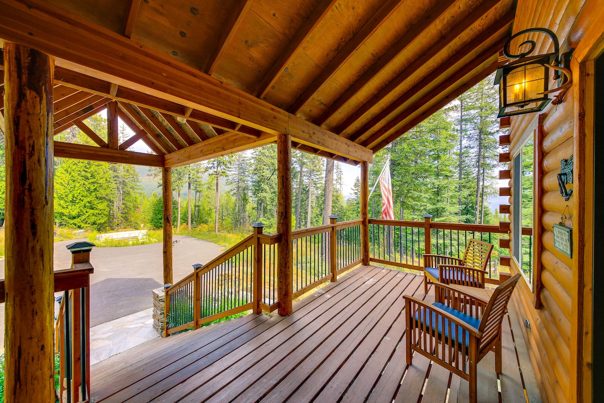 A spacious outdoor deck features wooden flooring and towering exposed beams. Two rocking chairs are positioned for outdoor seating, overlooking the forested surroundings. A flagpole can be seen in the distance, complementing the tranquil mountain atmosphere.