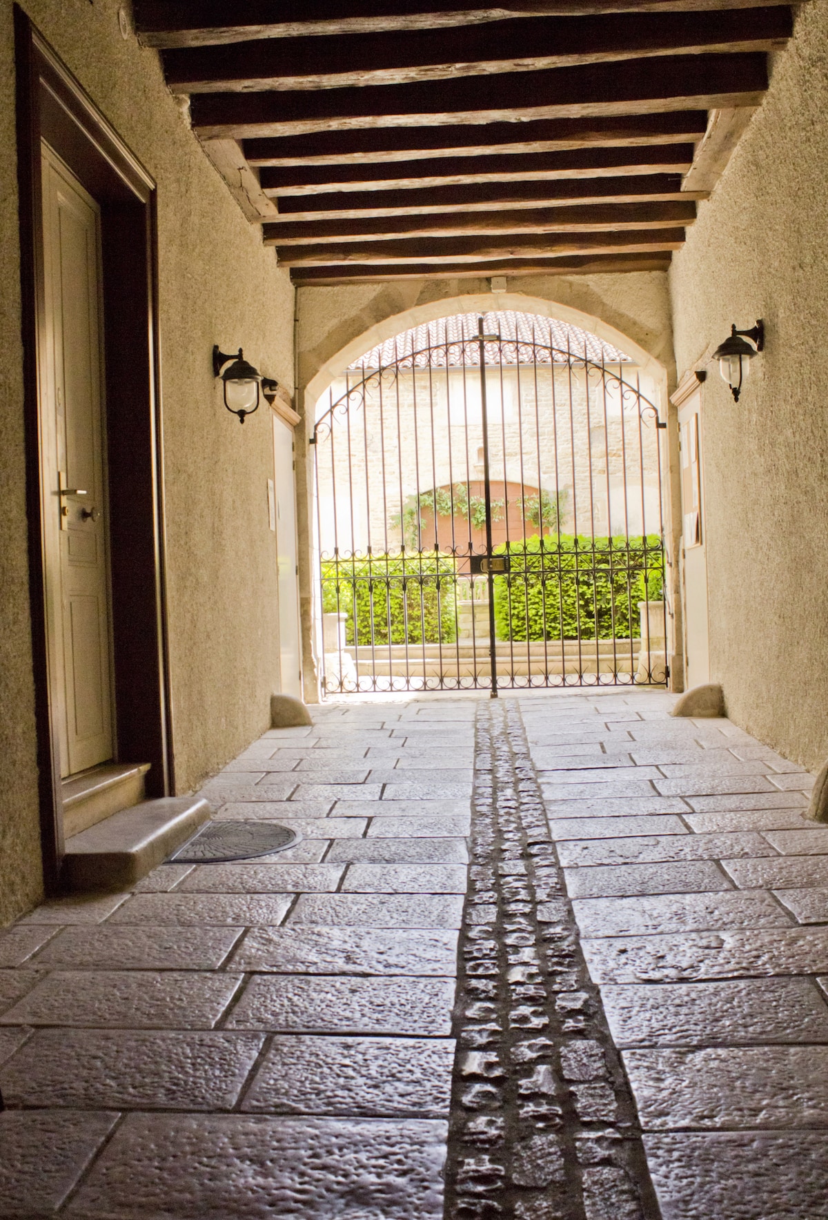 A stone pathway extends down a hallway, leading to an ornate gate framed by greenery. The hallway features an arched ceiling with wooden beams, and smooth walls with a light-colored finish. Two wall sconces provide soft illumination along the inviting corridor.