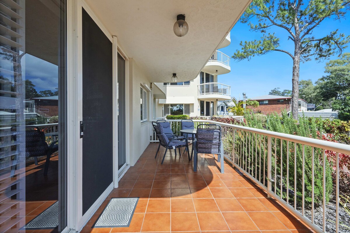 A spacious balcony is presented, featuring a tiled floor and surrounded by lush greenery. Two comfortable chairs and a small table create an inviting space for relaxation, while the nearby railing provides a clear view of the outdoor surroundings.