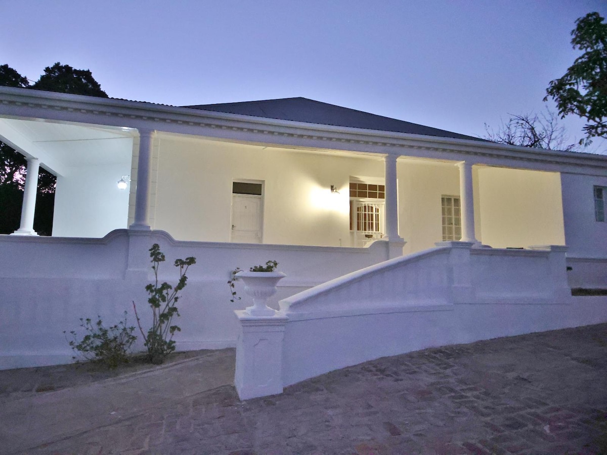 An elegant exterior showcases a white-painted structure with a wraparound porch supported by tall columns. Soft lighting highlights the entrance and the surrounding greenery, while a gently sloping pathway leads up to the inviting steps.