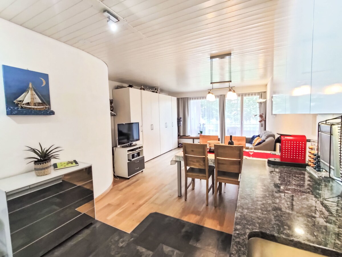 A spacious living and dining area features modern furnishings, including a dining table surrounded by four chairs. Natural light filters through large windows, highlighting a cozy seating arrangement with a sofa. A flat-screen TV and wall art complement the contemporary decor.