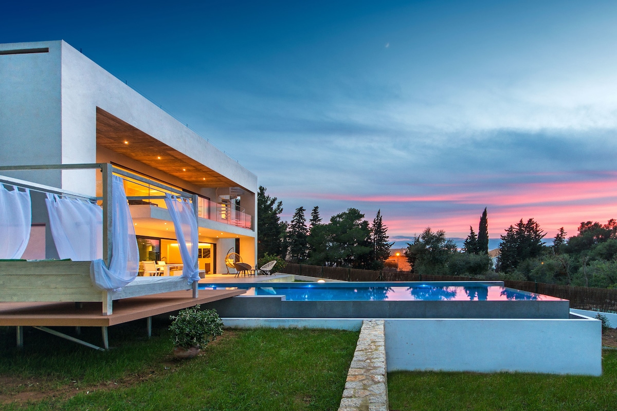 A modern villa is showcased against a sunset sky, featuring a large infinity pool that reflects the colors above. The spacious deck is adorned with sheer drapes, and comfortable outdoor seating is visible. Lush greenery surrounds the property, enhancing the serene atmosphere.