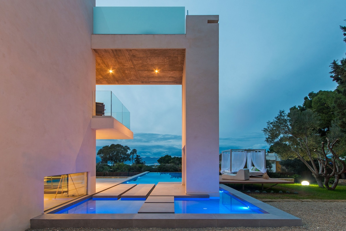 The villa's modern architecture is showcased with a cantilevered upper level overlooking an illuminated infinity pool. Soft blue lighting enhances the pool area, while a Balinese lounger and shaded space are visible in the background, providing a serene outdoor environment.